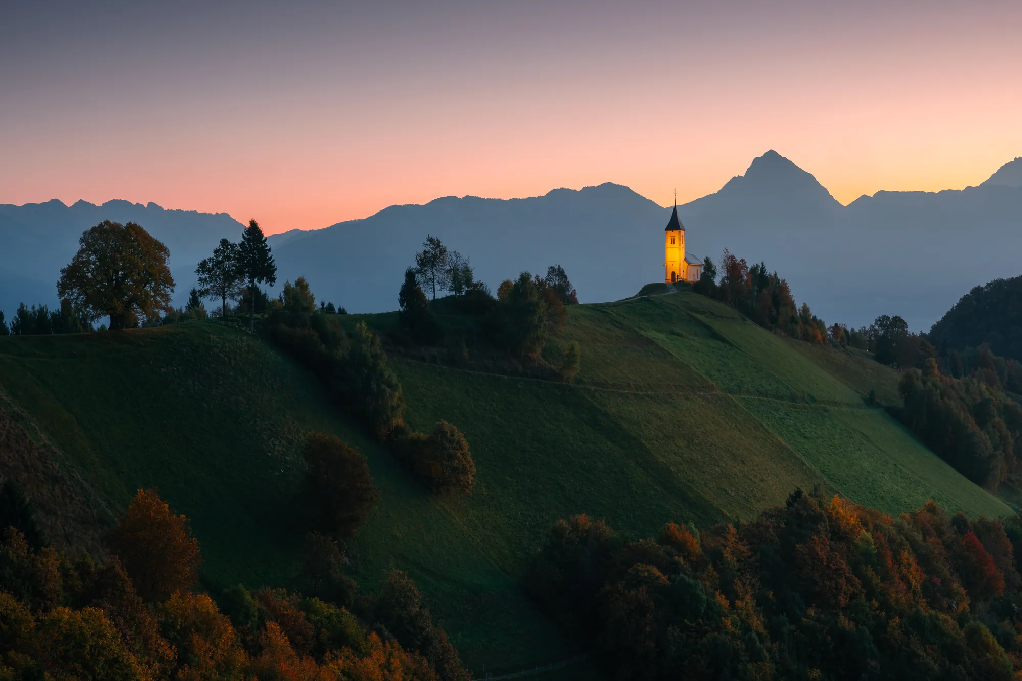 Kostel na kopci v Jamniku při východu slunce s alpskou siluetou, krajinářská fotografie, fotokurz Slovinsko